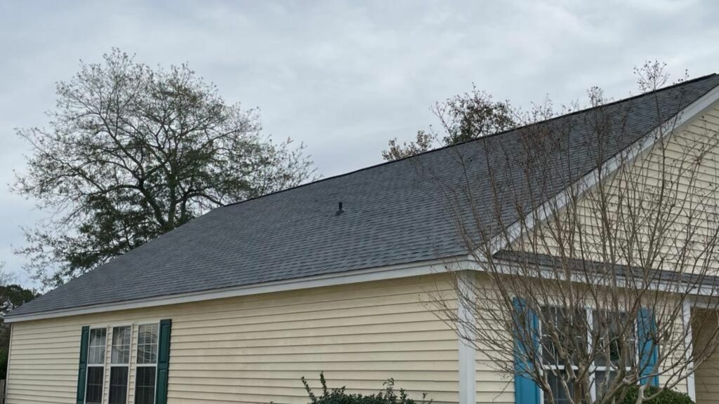 House with light yellow siding, dark gray roof, white trim, and mismatched green and blue shutters, shown on a cloudy day with bare trees.