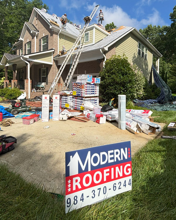 Residential roofing project in progress with workers on roof of two-story brick house, ladders, stacked shingles, equipment in yard, and Modern Roofing sign in foreground.