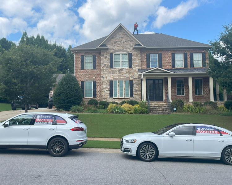 Two-story house with brick and stone exterior, person standing on roof, two white Modern Roofing vehicles parked on street, and landscaped yard under partly cloudy sky.