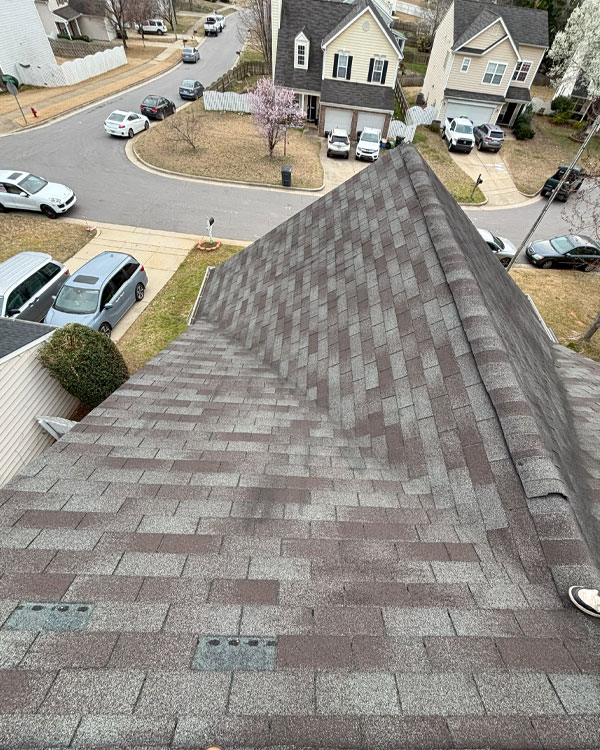 View from rooftop showing brown and gray asphalt shingles, ridge line, and slope, with suburban street, houses, cars, and blooming trees visible below.