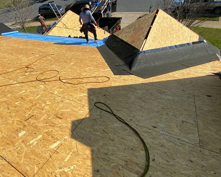 Residential roofing construction scene with OSB sheathing, worker in safety harness, dormers with black underlayment, blue tarp, and truck with materials nearby.