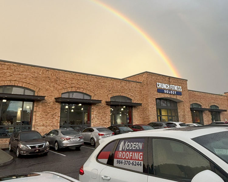Commercial building with Crunch Fitness Select sign, brick facade, parked cars, rainbow in sky, and white vehicle with Modern Roofing signage in foreground