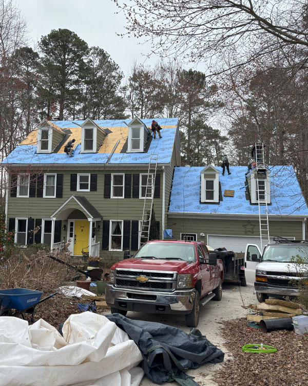 Two-story house with greenish-gray exterior and blue synthetic roof underlayment, five workers on roof, ladders, red and white pickup trucks in driveway, and wooded background.